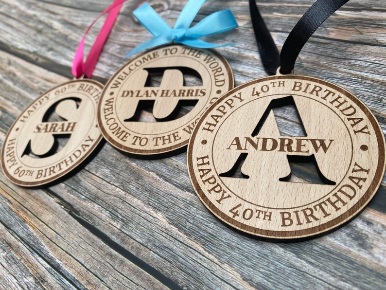 Three wooden decorations with a personalised initial and name. The first decoration is pink and says "Happy 60th Birthday Sarah". The second decoration is blue and says "Welcome to the World Dylan Harris". The third decoration is black and says "Happy 40th Birthday Andrew".