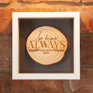 May include: A framed wood slice with the text "Be mine ALWAYS" and "ENGAGED 2021" engraved on it. The wood slice is in a white frame with a gray mat.