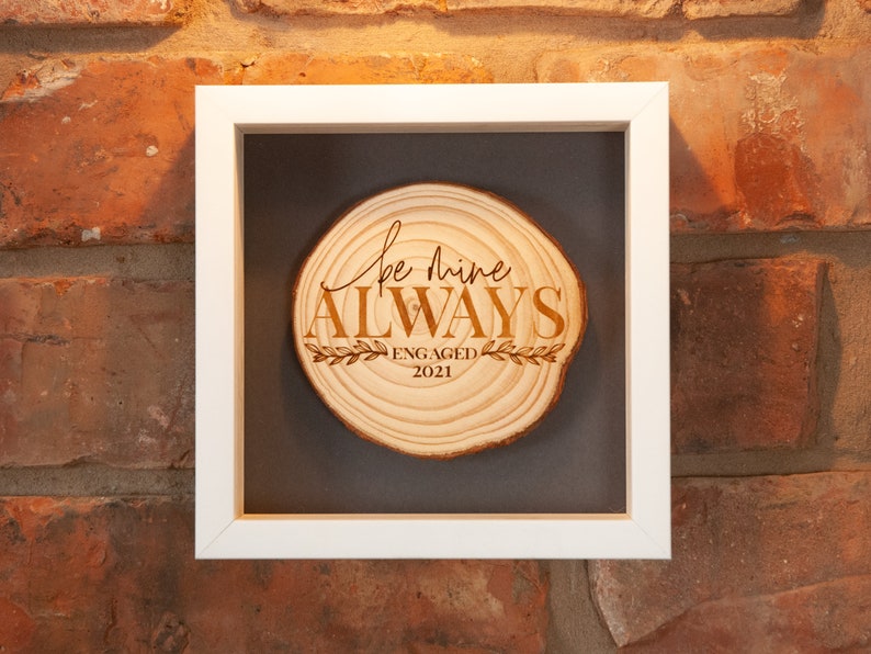 May include: A framed wood slice with the text "Be mine ALWAYS" and "ENGAGED 2021" engraved on it. The wood slice is in a white frame with a gray mat.