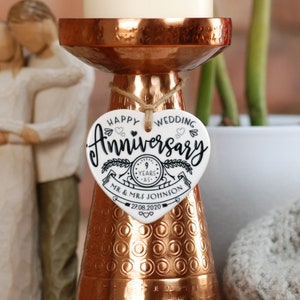 May include: White ceramic heart-shaped ornament with black text that reads "Happy Wedding Anniversary 9 Years - Mr. & Mrs. Johnson 27.08.2020". The ornament is hanging from a copper candle holder.