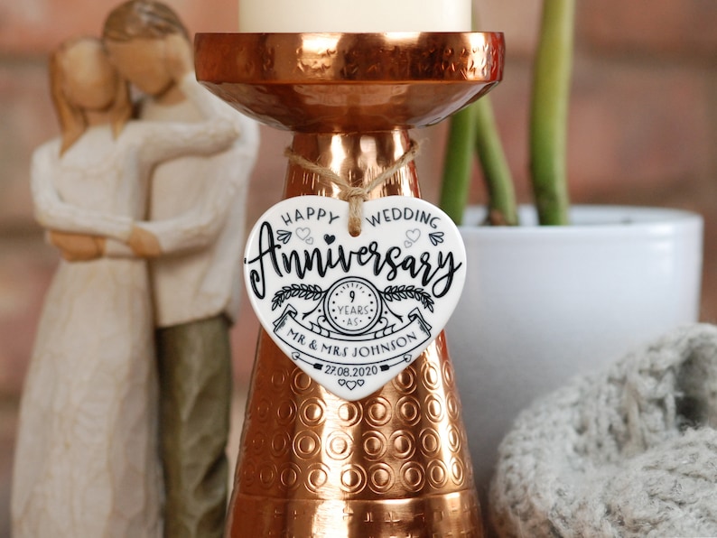 May include: White ceramic heart-shaped ornament with black text that reads "Happy Wedding Anniversary 9 Years - Mr. & Mrs. Johnson 27.08.2020". The ornament is hanging from a copper candle holder.