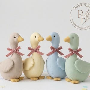 May include: Four plush goose toys in beige, yellow, light blue, and sage green. Each goose features a yellow beak, small black eyes, and a dusty rose velvet ribbon tied around its neck. The logo in the top right corner reads Pour Filles.