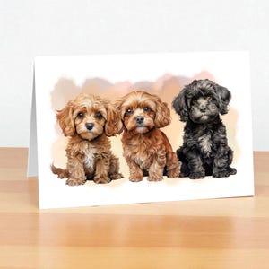 Può includere: Un biglietto di auguri con tre adorabili cuccioli. I cuccioli hanno una soffice pelliccia nei toni del marrone e del nero, con occhi espressivi. Lo sfondo presenta colori tenui e sfumati di pesca e beige. Il biglietto è appoggiato su una superficie di legno.