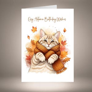 Può includere: Un biglietto d'auguri bianco con un'illustrazione ad acquerello di un gatto soriano marrone chiaro che indossa una sciarpa in maglia arancione. Il gatto è circondato da foglie autunnali arancioni e gialle. Il testo recita Cosy Autumn Birthday Wishes.