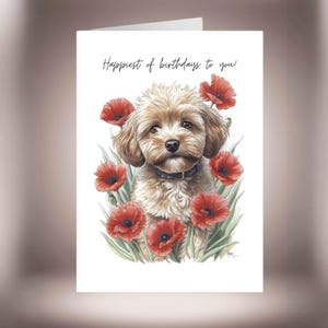Può includere: Biglietto di auguri con un'illustrazione ad acquerello di un cane marrone chiaro circondato da papaveri rossi e foglie verdi. Il biglietto recita "Happiest of birthdays to you!" in una scrittura elegante.