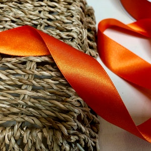 May include: A close-up of a bright orange satin ribbon. The ribbon is lying on a woven surface.