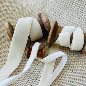 May include: Ivory velvet ribbon on wooden spools. The ribbon is unwound from the spools, which have a natural wood finish. The background is a textured, neutral-toned fabric. The ribbon has a soft, luxurious appearance, ideal for crafts and embellishments.