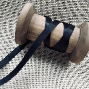 Pu&ograve; includere: Un rocchetto di nastro di raso nero avvolto attorno a un rocchetto di legno. Il nastro &egrave; liscio e lucido.