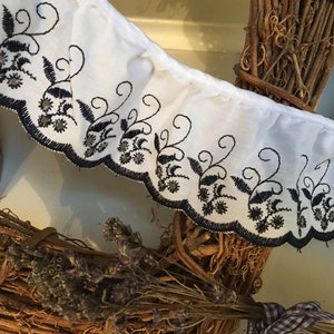 May include: White fabric trim with a scalloped edge, embroidered with black floral and scroll designs. The trim is ruffled and appears to be part of a larger textile. Decorative elements include small, gold-coloured accents.