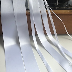 May include: A collection of white satin ribbon spools, arranged in a fan shape on a white surface. The ribbon is smooth and shiny, with a slight sheen.