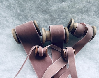 May include: Four spools of dusty rose velvet ribbon with wooden spools. The ribbon is arranged in a loose pile with some of the ribbon unraveled.