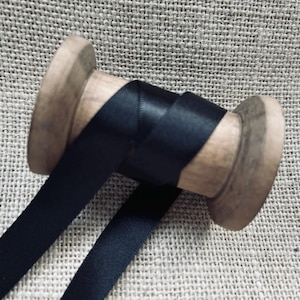 Pu&ograve; includere: Un rocchetto di nastro di raso nero avvolto attorno a un rocchetto di legno. Il nastro &egrave; liscio e lucido.