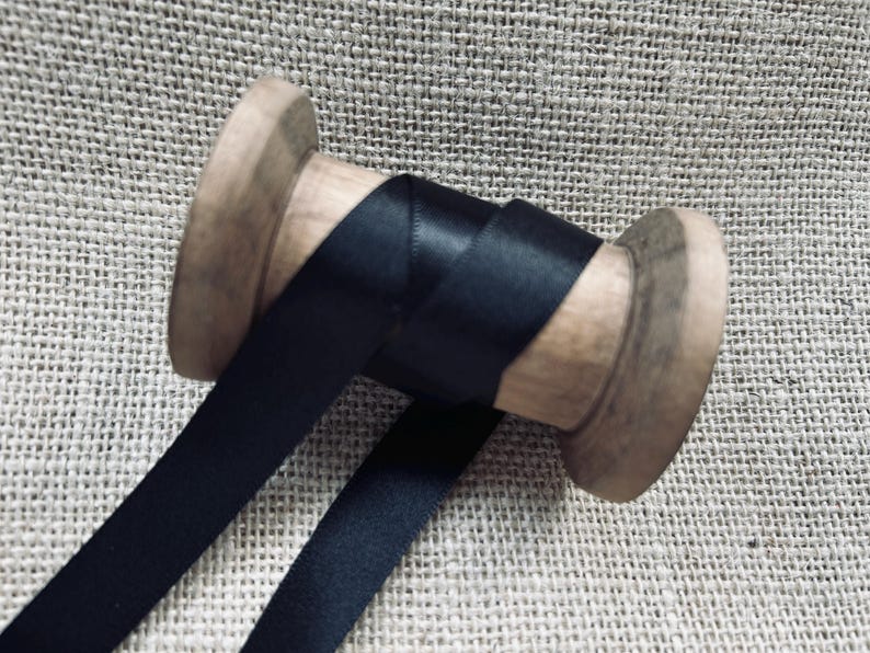Pu&ograve; includere: Un rocchetto di nastro di raso nero avvolto attorno a un rocchetto di legno. Il nastro &egrave; liscio e lucido.
