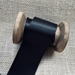 Pu&ograve; includere: Un rocchetto di nastro di raso nero avvolto attorno a un rocchetto di legno. Il nastro &egrave; liscio e lucido.