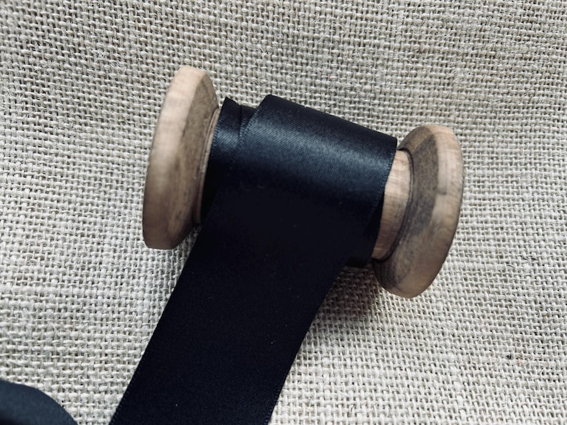 Pu&ograve; includere: Un rocchetto di nastro di raso nero avvolto attorno a un rocchetto di legno. Il nastro &egrave; liscio e lucido.