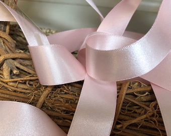 May include: Pink satin ribbon with a smooth, shiny finish. The ribbon is folded and arranged on a brown, woven surface.