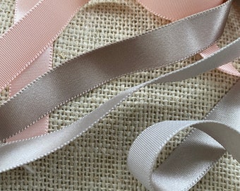 May include: A close-up of three different colored ribbons on a burlap background. The ribbons are a light pink, a light gray, and a white color. The ribbons are arranged in a criss-cross pattern.