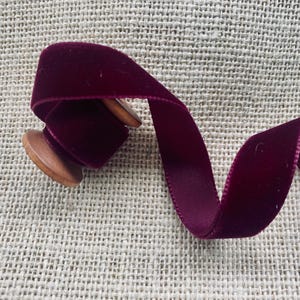 May include: A spool of deep burgundy velvet ribbon. The ribbon is wound around a wooden spool and is lying on a burlap surface.