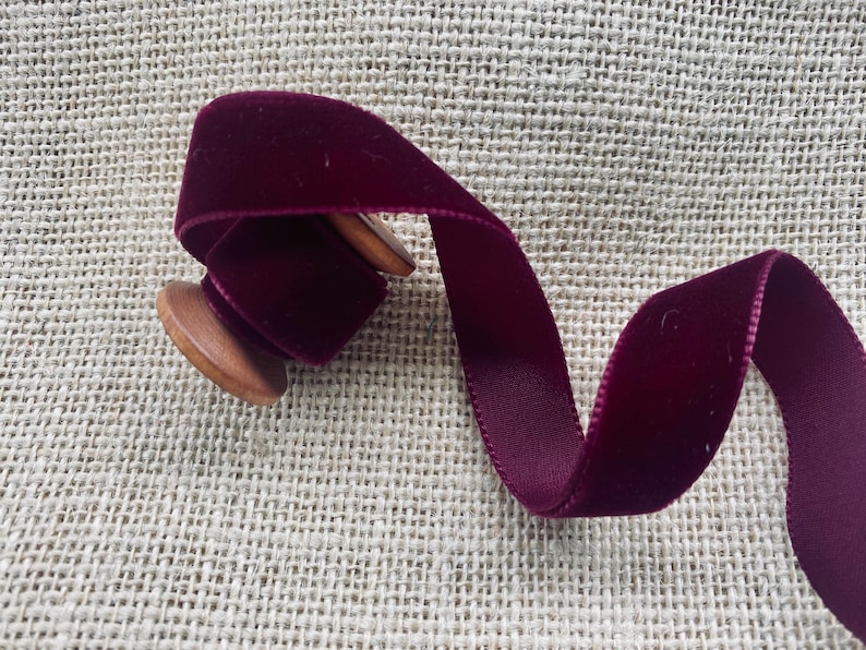 May include: A spool of deep burgundy velvet ribbon. The ribbon is wound around a wooden spool and is lying on a burlap surface.