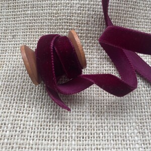 May include: A spool of burgundy velvet ribbon with a wooden spool. The ribbon is wound around the spool and is ready to be used for crafting or decorating.