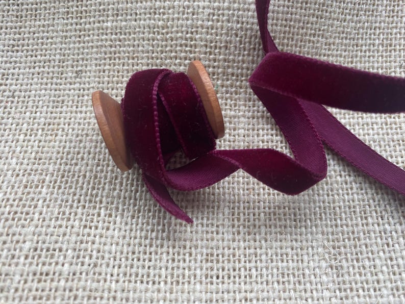 May include: A spool of burgundy velvet ribbon with a wooden spool. The ribbon is wound around the spool and is ready to be used for crafting or decorating.