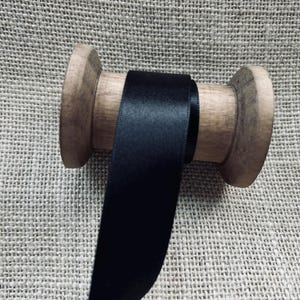 Pu&ograve; includere: Un rotolo di nastro di raso nero avvolto attorno a un rocchetto di legno. Il nastro &egrave; liscio e lucido.