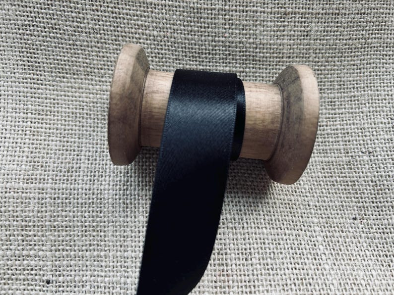 Pu&ograve; includere: Un rotolo di nastro di raso nero avvolto attorno a un rocchetto di legno. Il nastro &egrave; liscio e lucido.