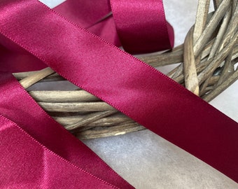 May include: Close-up of a deep burgundy satin ribbon. The ribbon is folded and woven through a wicker wreath.