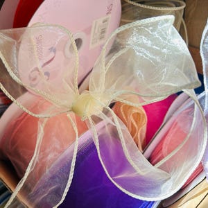 Peut inclure: Gros plan sur une boîte remplie de rubans colorés. Un nœud en ruban transparent crème est au premier plan. Des rouleaux de rubans roses, violets et oranges sont visibles. Les rubans sont de différentes matières et textures.