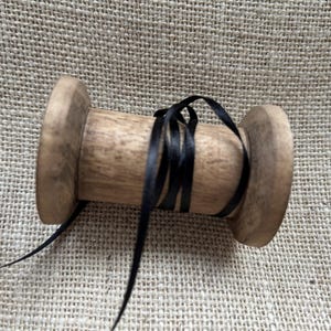 Pu&ograve; includere: Un rocchetto di legno con un nastro di raso nero avvolto attorno. Il rocchetto &egrave; su una superficie di juta.