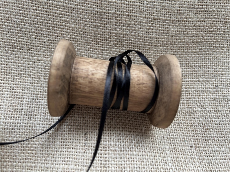 Pu&ograve; includere: Un rocchetto di legno con un nastro di raso nero avvolto attorno. Il rocchetto &egrave; su una superficie di juta.