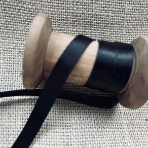 Pu&ograve; includere: Un rocchetto di nastro di raso nero avvolto attorno a un rocchetto di legno. Il nastro &egrave; liscio e lucido.