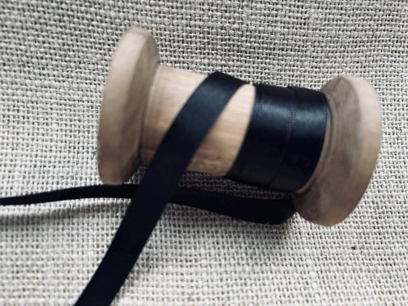Pu&ograve; includere: Un rocchetto di nastro di raso nero avvolto attorno a un rocchetto di legno. Il nastro &egrave; liscio e lucido.