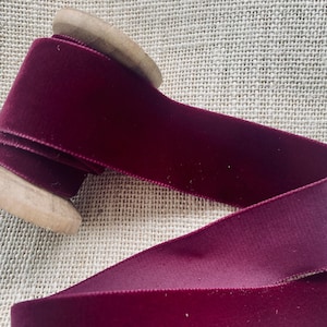 May include: A spool of deep burgundy velvet ribbon wound around a wooden spool. The ribbon is smooth and has a subtle sheen.