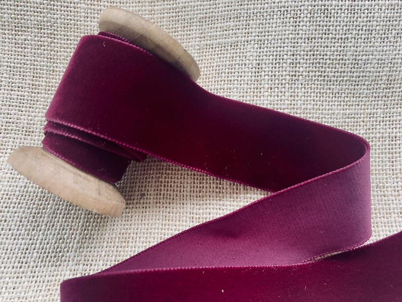 May include: A spool of deep burgundy velvet ribbon wound around a wooden spool. The ribbon is smooth and has a subtle sheen.