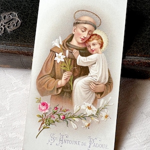 May include: A rectangular religious prayer card featuring a color illustration of Saint Anthony of Padua holding the Christ Child. The saint wears a brown habit and holds white lilies. The text St. Antoine de Padoue is printed at the bottom.