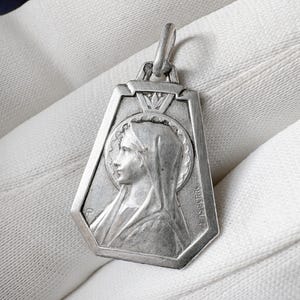 Puede incluir: Un colgante de plata que representa el perfil de una mujer con halo, probablemente un icono religioso. El colgante tiene una forma geométrica y un lazo para una cadena. El metal tiene un aspecto ligeramente envejecido, con detalles finos.