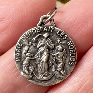 May include: A silver-toned pendant with the words "MARIE QUI DEFAIT LES NOEUDS" encircling a central image. The image depicts a robed figure with several smaller figures. The pendant has a loop for a chain.