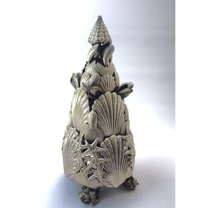 May include: A silver-colored metal sculpture of a tree made from seashells. The tree is cone-shaped and has a base of three small seashells. The tree is decorated with various seashells, including starfish.
