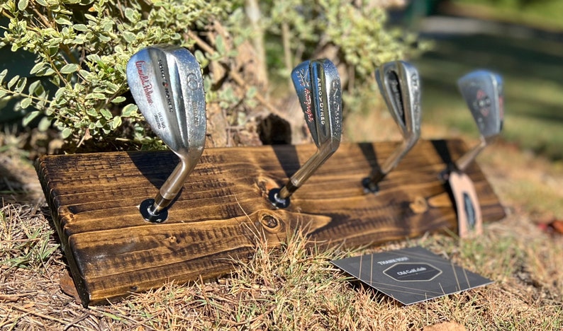 May include: A wooden rack with four golf club heads mounted as hooks. The rack is stained dark brown and has a rustic look. The golf clubs are silver and have various brand names.