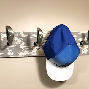 May include: A rustic wooden coat rack with three golf club heads as hooks. A blue and white baseball cap hangs on one of the hooks.