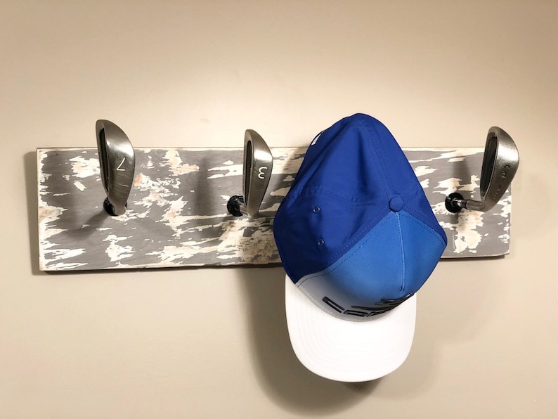 May include: A rustic wooden coat rack with three golf club heads as hooks. A blue and white baseball cap hangs on one of the hooks.