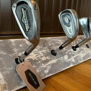 May include: A rustic wooden rack with four vintage golf clubs hanging from it. The rack is painted gray with white distressing. A tag with the text "Old Golfclub" is attached to one of the clubs.