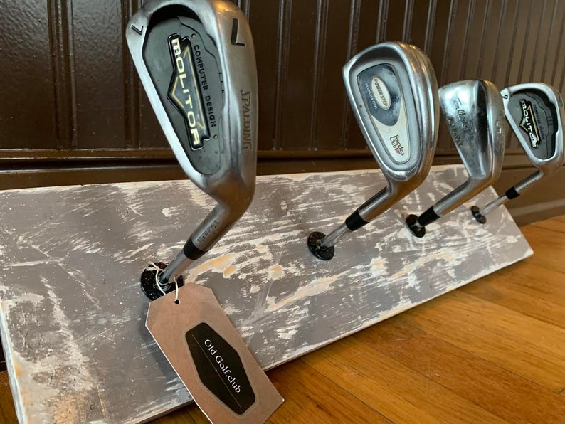 May include: A rustic wooden rack with four vintage golf clubs hanging from it. The rack is painted gray with white distressing. A tag with the text "Old Golfclub" is attached to one of the clubs.