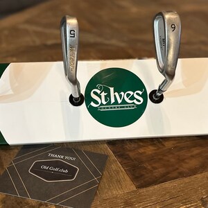 May include: A wooden golf club rack with a white and green paint scheme. The rack has four slots for golf clubs and features the text "St Ives Country Club" in a green circle. A small brown tag with the text "Old Golf Club" is attached to the rack.