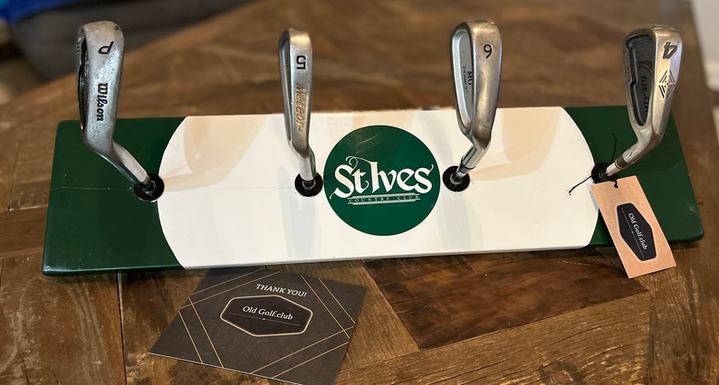 May include: A wooden golf club rack with a white and green paint scheme. The rack has four slots for golf clubs and features the text "St Ives Country Club" in a green circle. A small brown tag with the text "Old Golf Club" is attached to the rack.