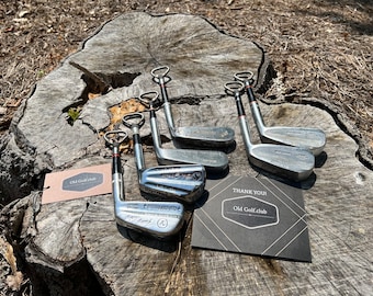 Könnte beinhalten: Sechs silberne Golfclub-Flaschenöffner auf einem Baumstumpf. Die Flaschenöffner sind in Form von Golfclubs gestaltet und haben einen Flaschenöffner am Ende. Der Baumstumpf ist braun und hat eine raue Textur. Auf dem Baumstumpf befindet sich eine kleine braune Karte mit dem Text "Old Golf club" und eine schwarze Karte mit dem Text "THANK YOU! Old Golf.club".