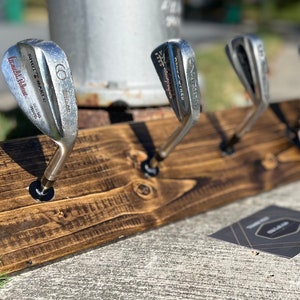 May include: A wooden golf club rack with four golf clubs hanging from it. The rack is stained dark brown and has a rustic look. The clubs are all silver and have different brands and numbers on them.