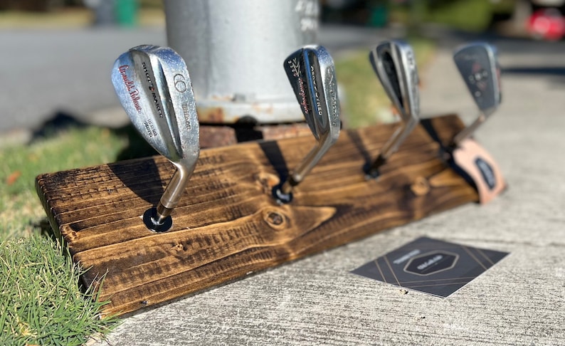 May include: A wooden golf club rack with four golf clubs hanging from it. The rack is stained dark brown and has a rustic look. The clubs are all silver and have different brands and numbers on them.