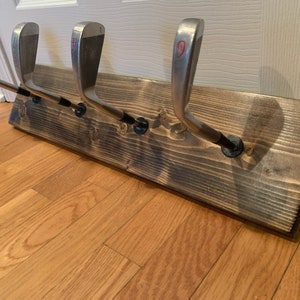 May include: A wooden wall-mounted coat rack with five golf club hooks. The rack is stained dark brown and has a rustic look.
